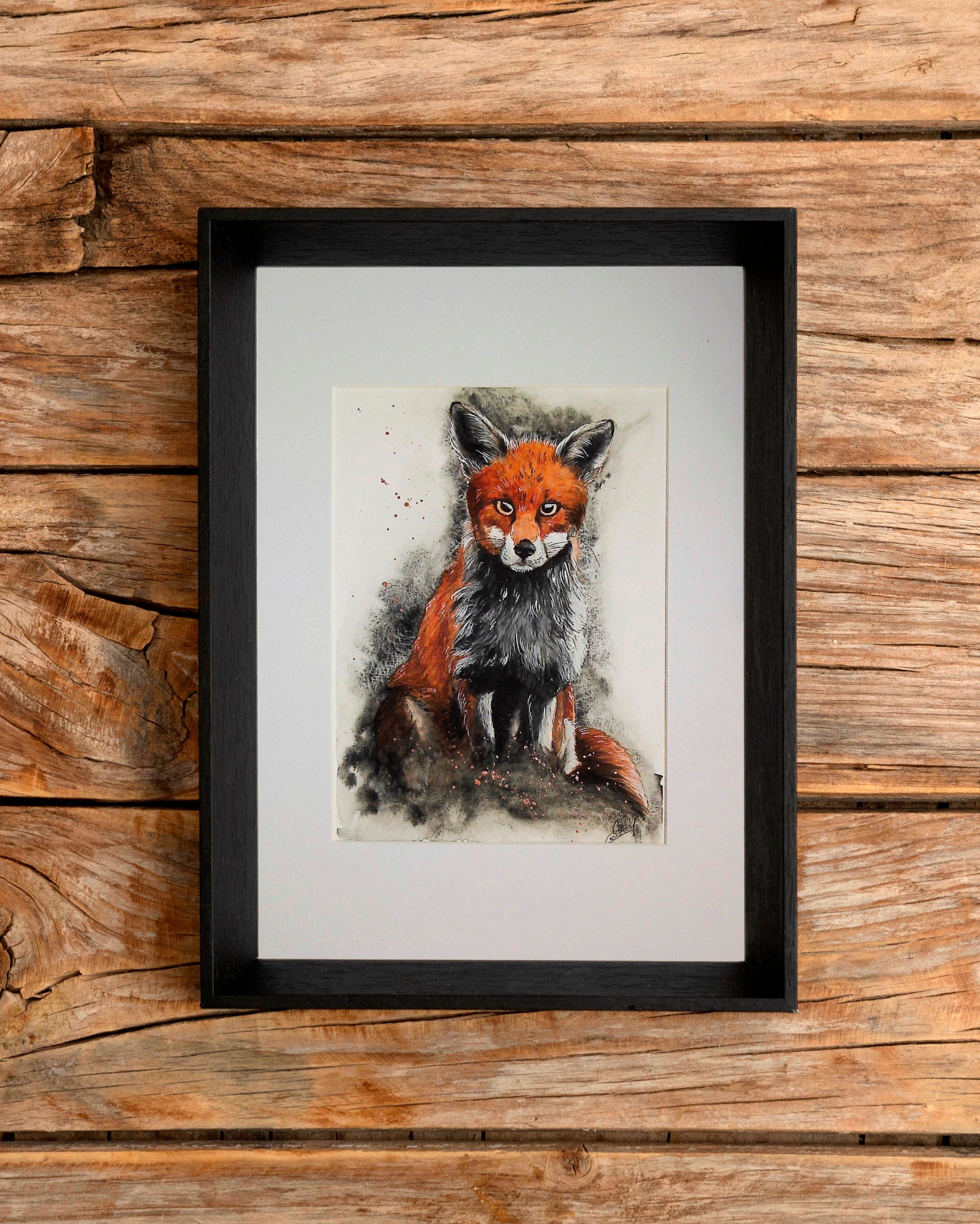 Another version of the watercolour fox painting in a frame on an wooden surface.