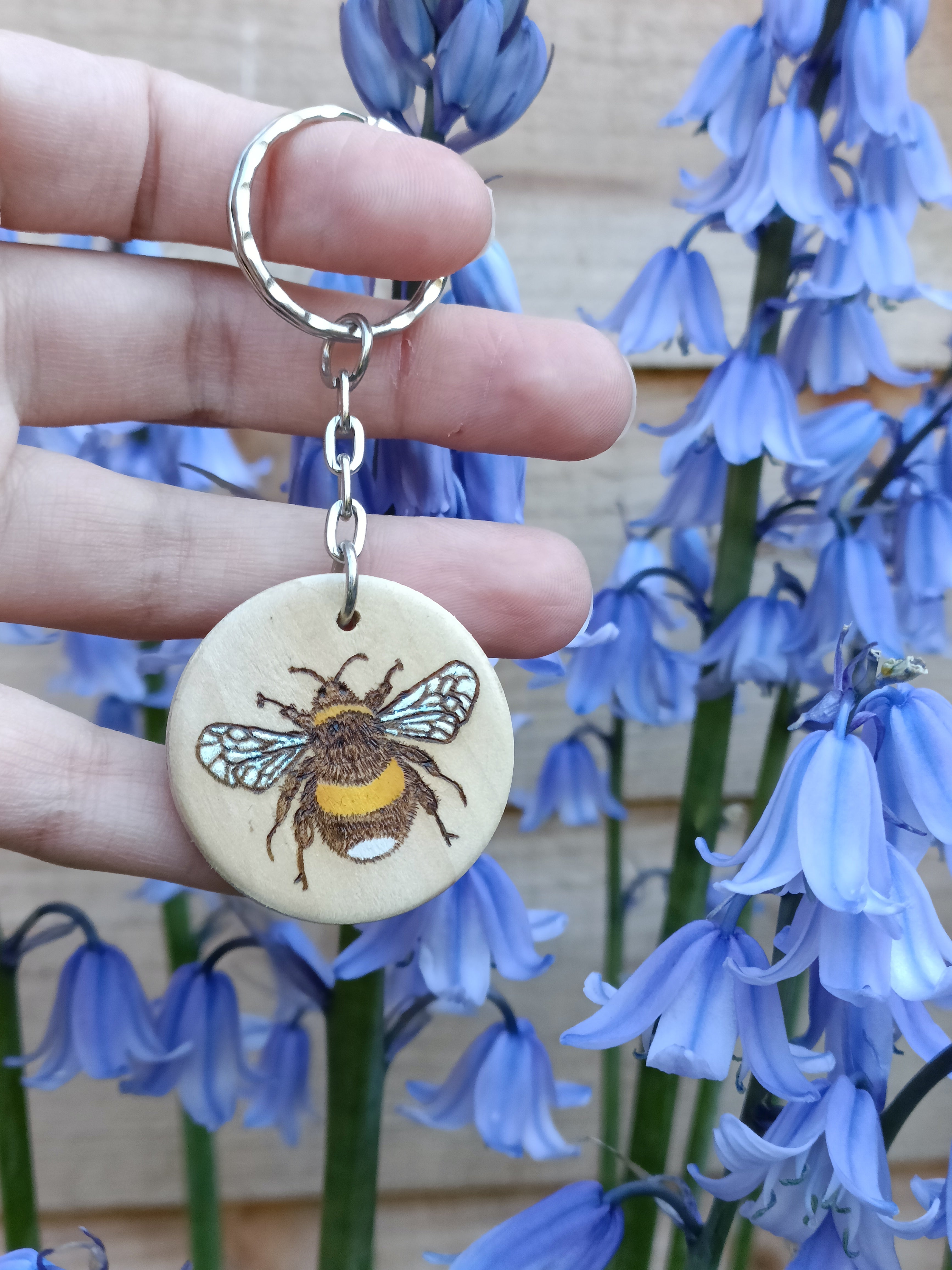 Hand holding a round wooden keyring with a woodburned and painted bumblebee illustration. Shown outside in natural daylight.