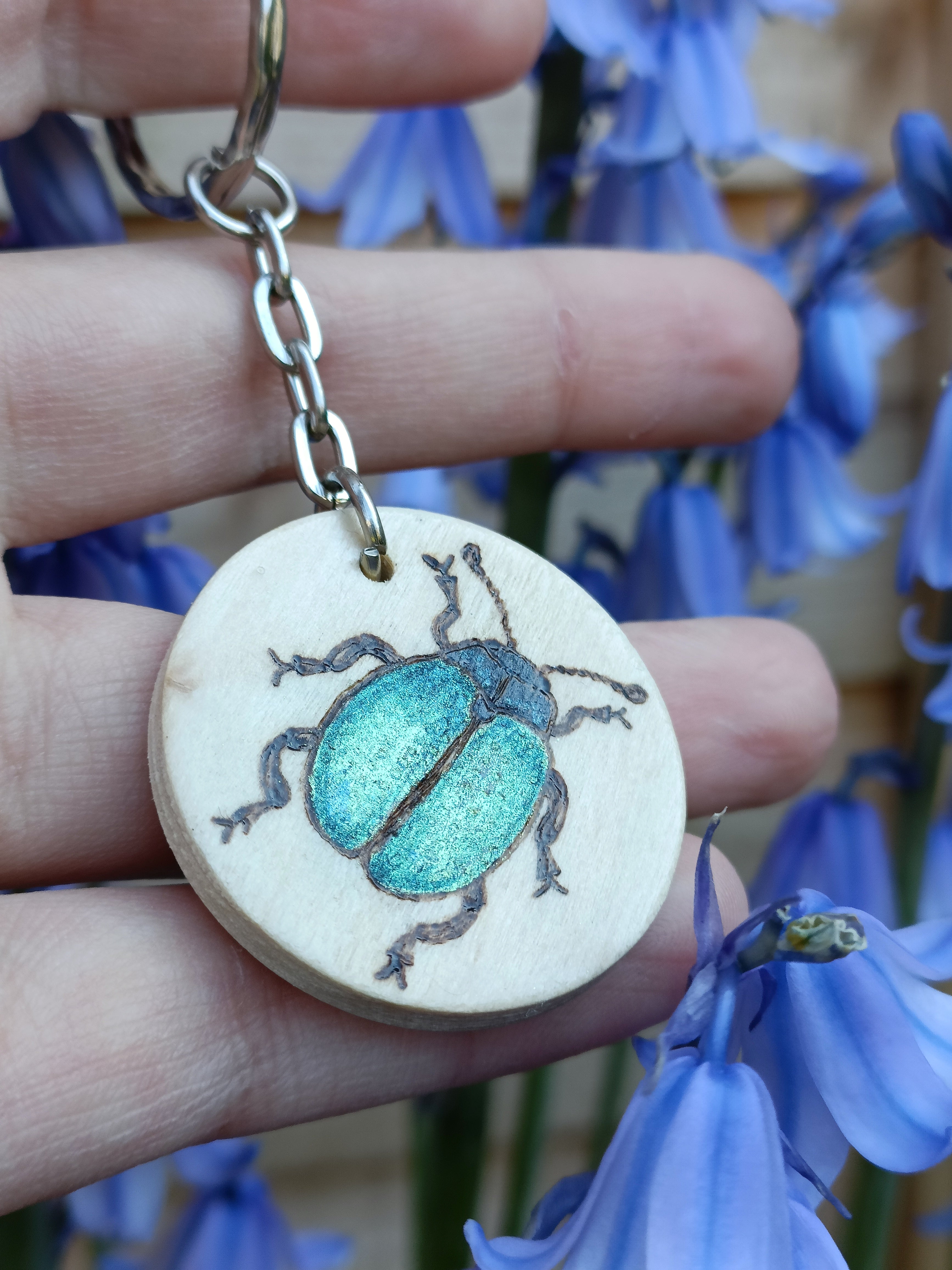 Hand holding a round wooden keyring with a blue beetle design that's woodburned and painted by hand. It's shown outside in natural daylight.
