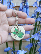 Hand holding a handmade keychain with a woodburned and painted walking leaf insect design, shown outside in natural light.