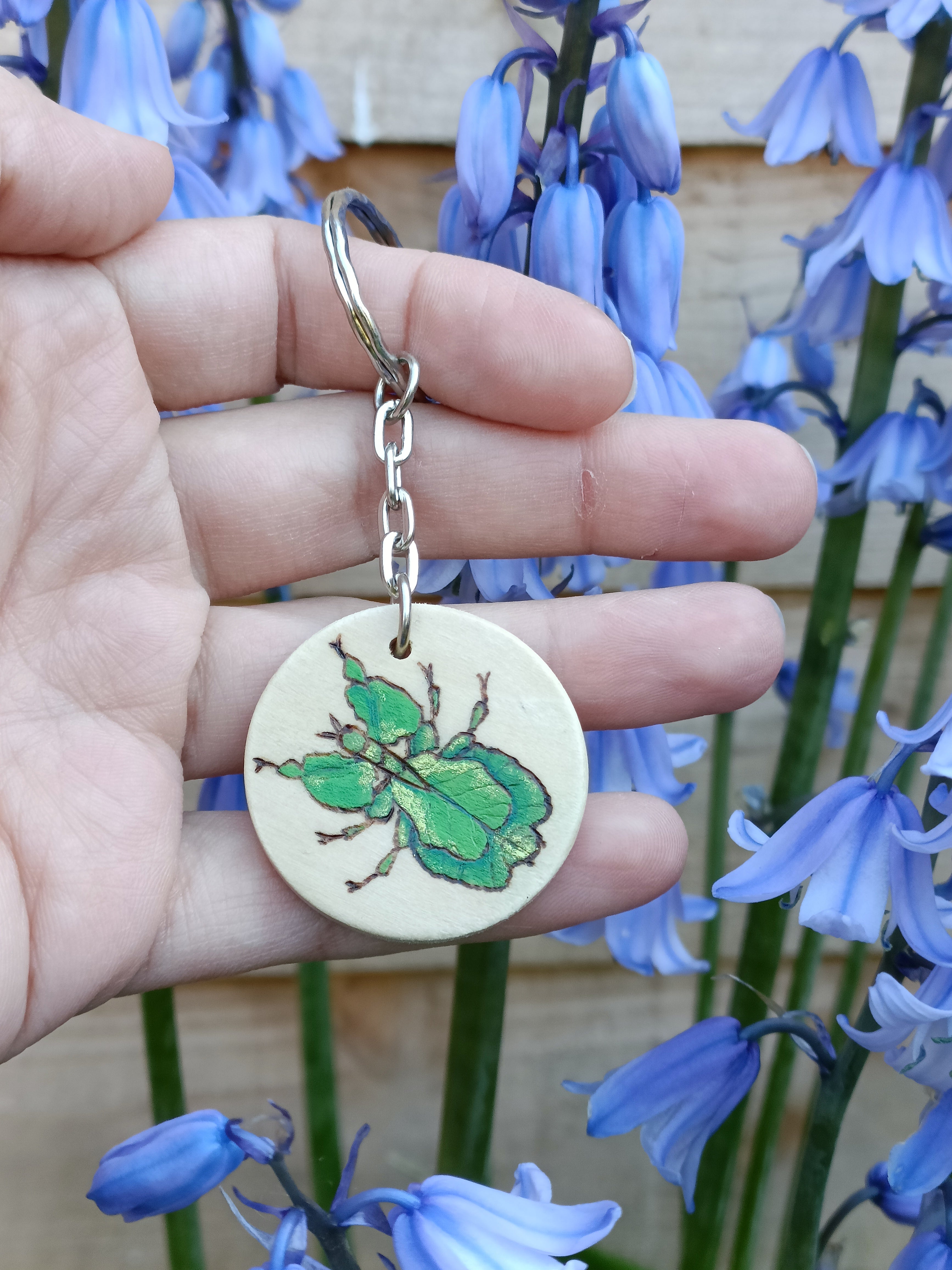 Hand holding a handmade keychain with a woodburned and painted walking leaf insect design, shown outside in natural light.