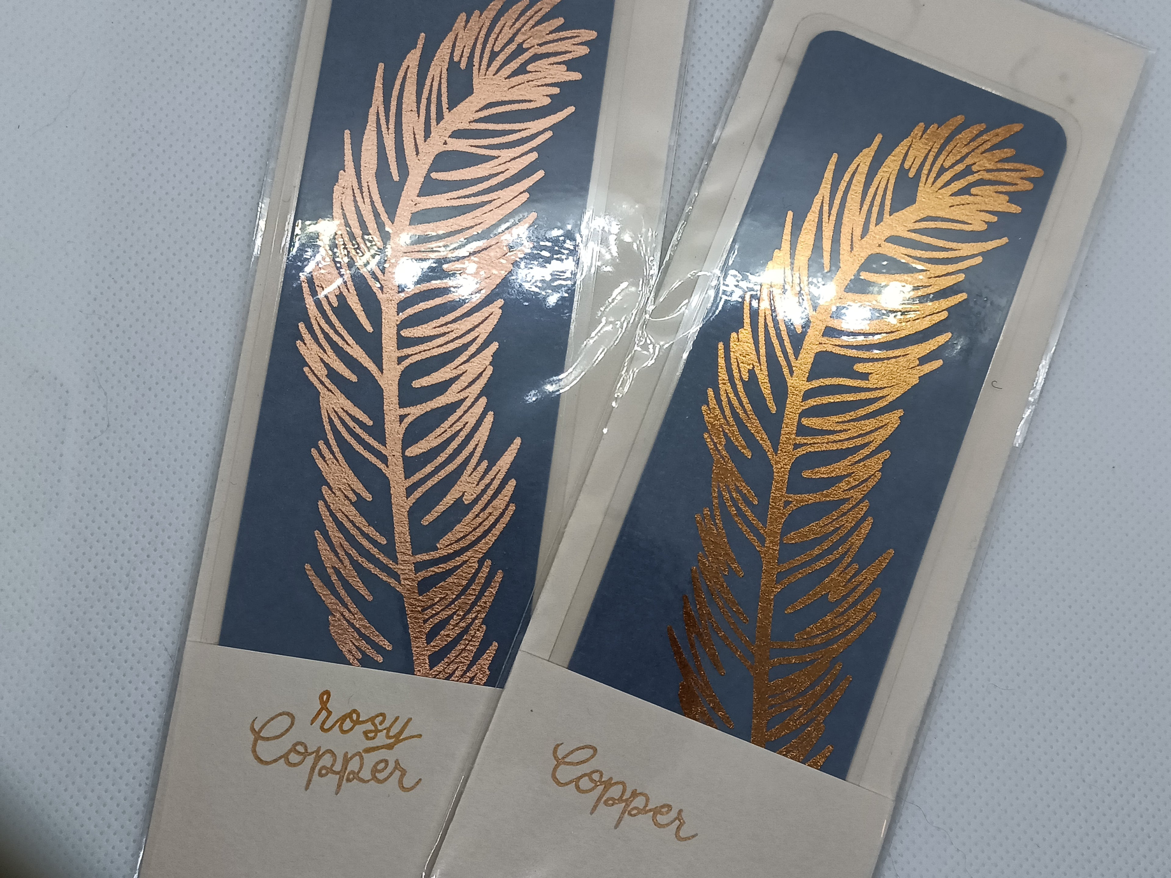 Two handfoiled metallic feather bookmarks next to each other, with 'rosy copper' and 'copper' text on their packaging, showing the difference between the two copper colour options in detail.
