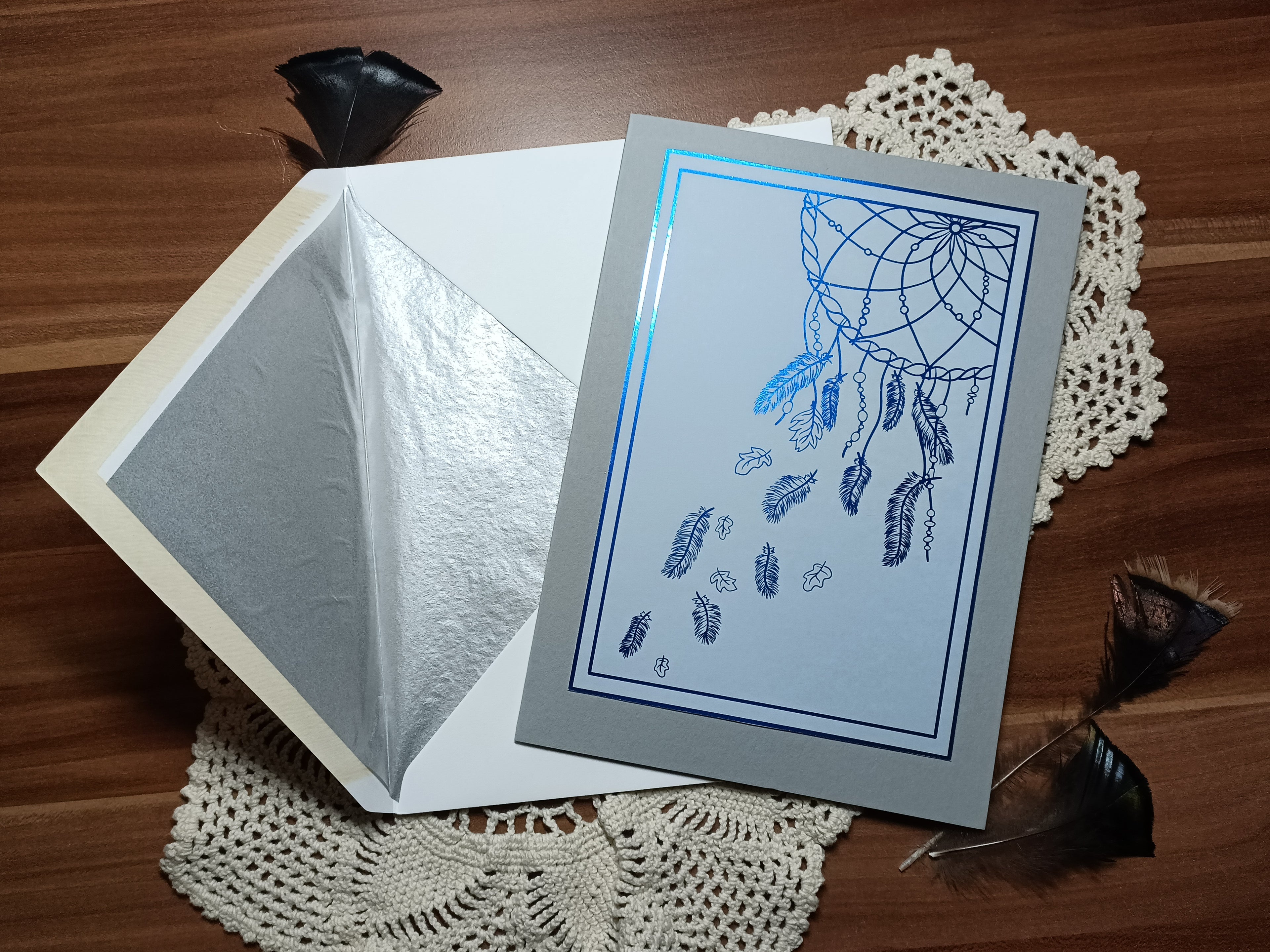 Handmade A5 greeting card with a dreamcatcher designed and foiled by hand, next to the accompanying envelope which features a silver inlay. The card is light grey and blue, the foil is a medium blue.