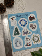 Sheet of Christmas-themed cat stickers with winter accessories, trees, and snowmen on a blue background, next to a ruler for scale.