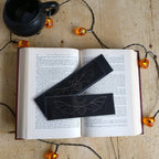 Open book with two bat-themed black bookmarks on a wooden surface with Halloween decor.
