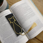 Rectangle-shaped black bookmark with a gold foiled cat outline with a tassel at the bottom, laid out on an open book.