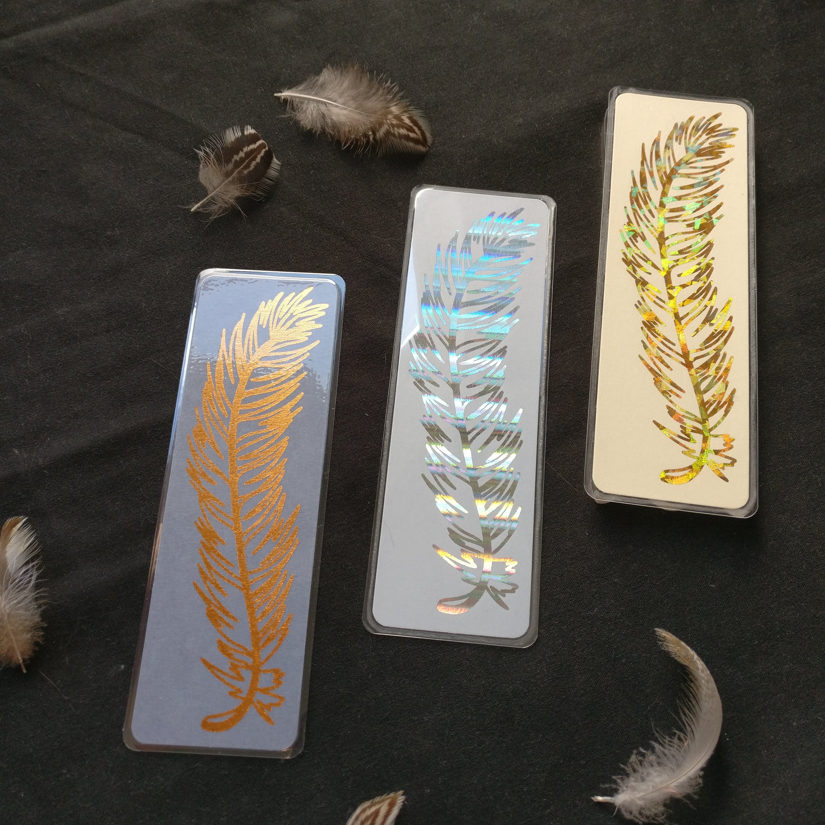 The three Feather Bookmark colour options, from left to right: copper, holographic silver, and holographic (crystal) gold.