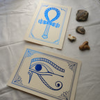 Two foil prints, blue foil on cream card, with decorative stones. The prints depict Egyptian hieroglyphs, the top one is of the Ankh symbol, the bottom one of the Eye of Horus.