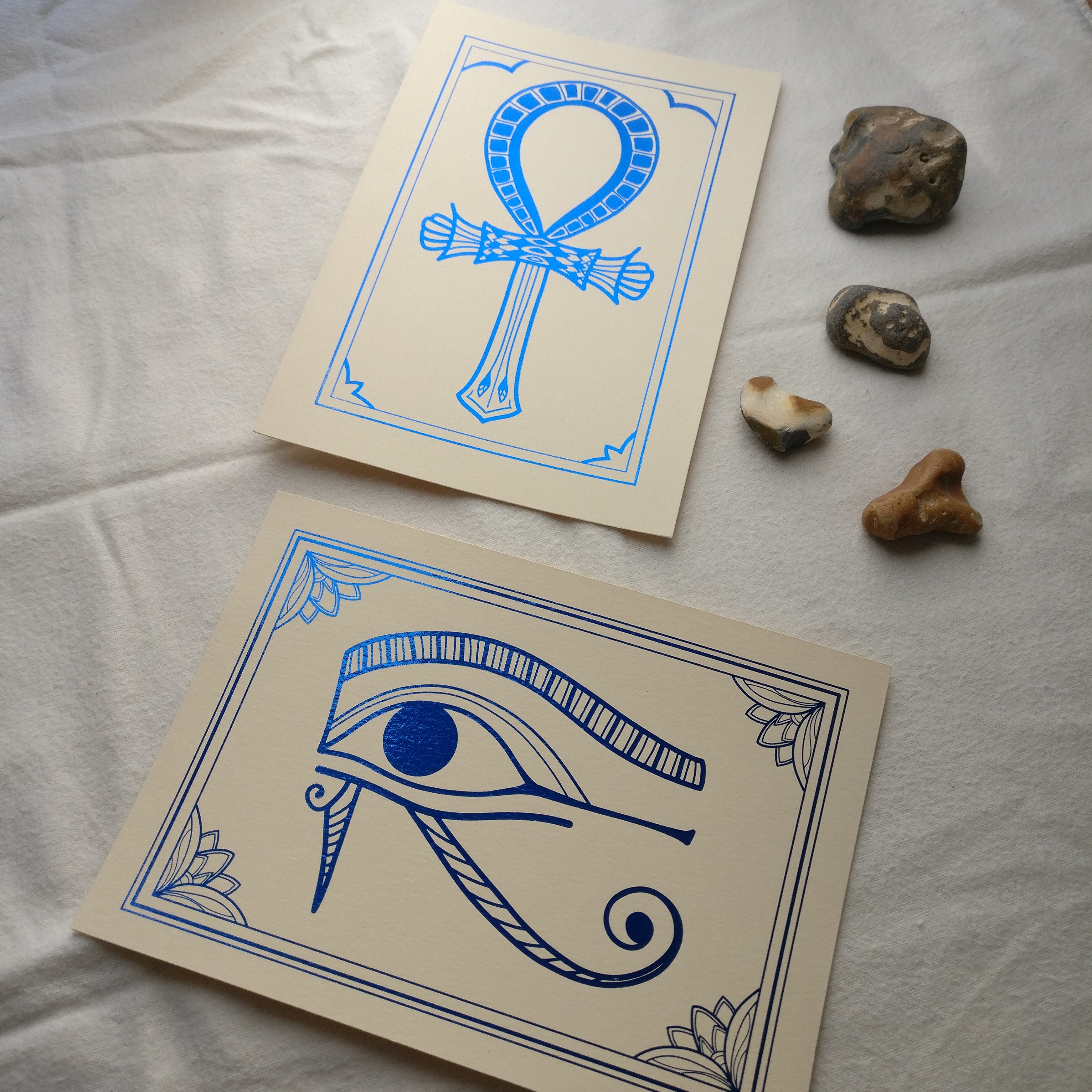 Two foil prints, blue foil on cream card, with decorative stones. The prints depict Egyptian hieroglyphs, the top one is of the Ankh symbol, the bottom one of the Eye of Horus.