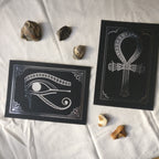 Two foil prints, silver foil on black card, with decorative stones. The prints depict Egyptian hieroglyphs, the left one is of the Eye of Horus, the right one of the Ankh symbol.