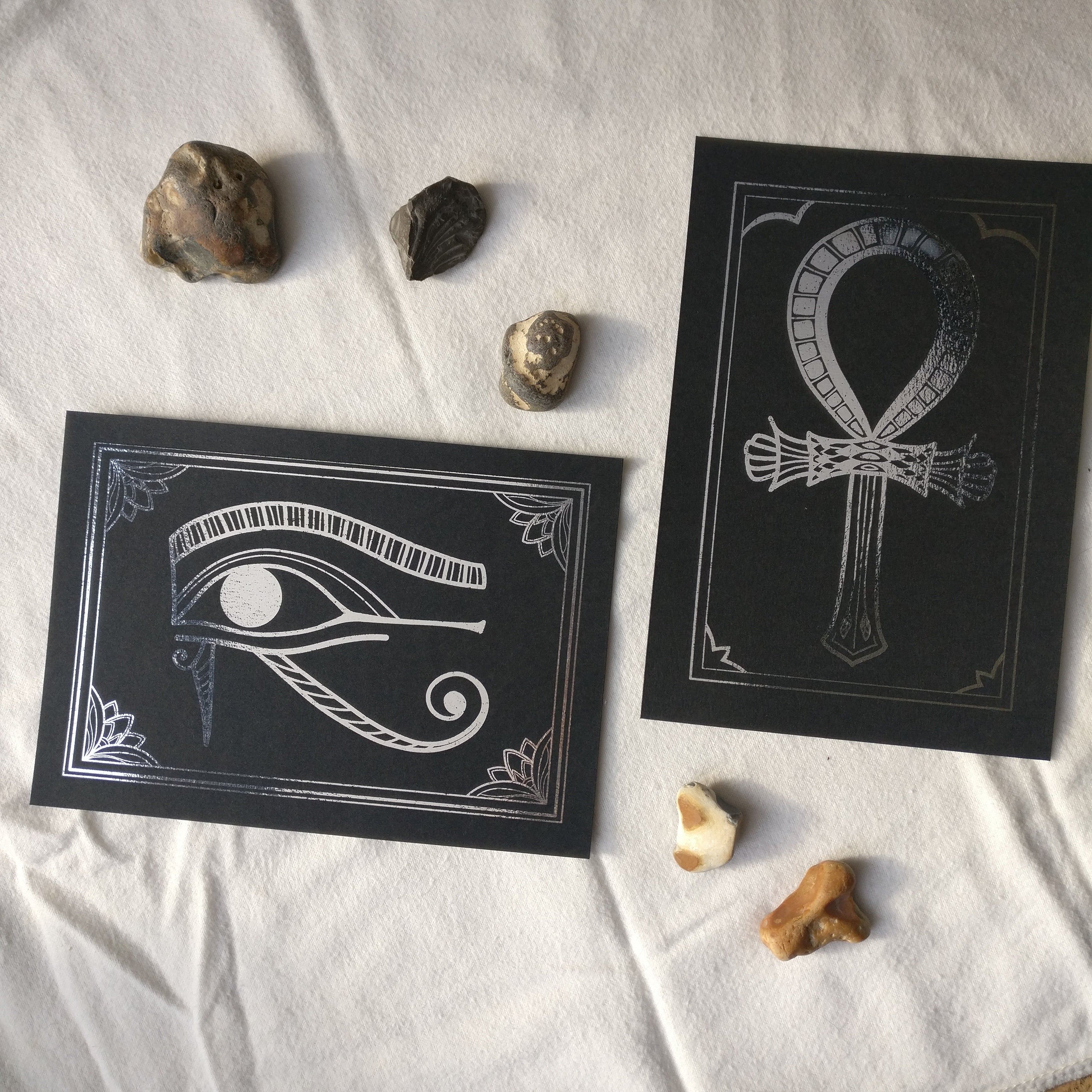 Two foil prints, silver foil on black card, with decorative stones. The prints depict Egyptian hieroglyphs, the left one is of the Eye of Horus, the right one of the Ankh symbol.