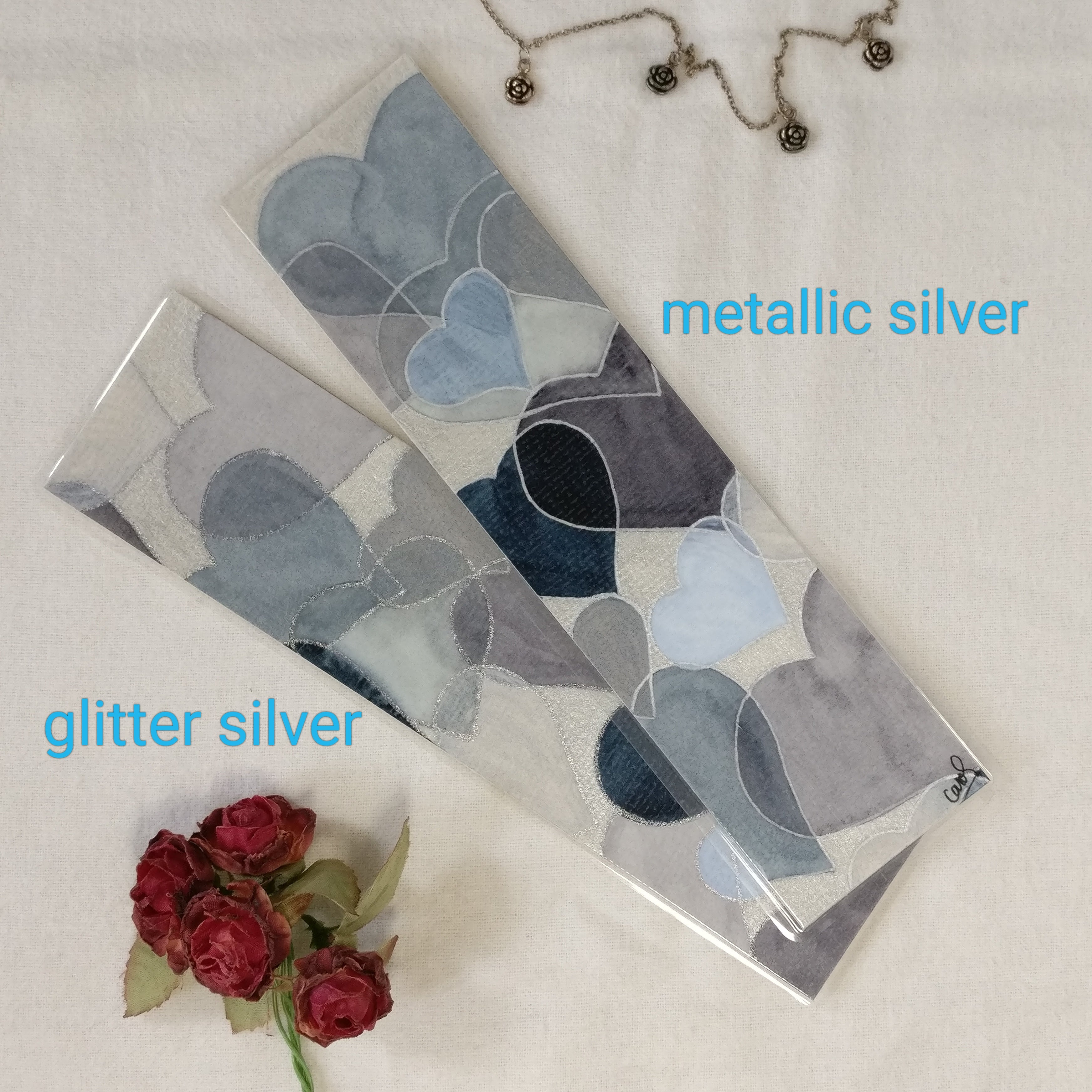 Two hand-painted watercolour hearts bookmarks in grey, one with glitter silver outlines and the other one with metallic silver outlines. The writing next to each bookmark makes clear which is which.