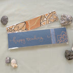 An orange Mandala Bookmark with one of the handmade package backings with a hand-written "happy reading" and a mandala corner in copper.