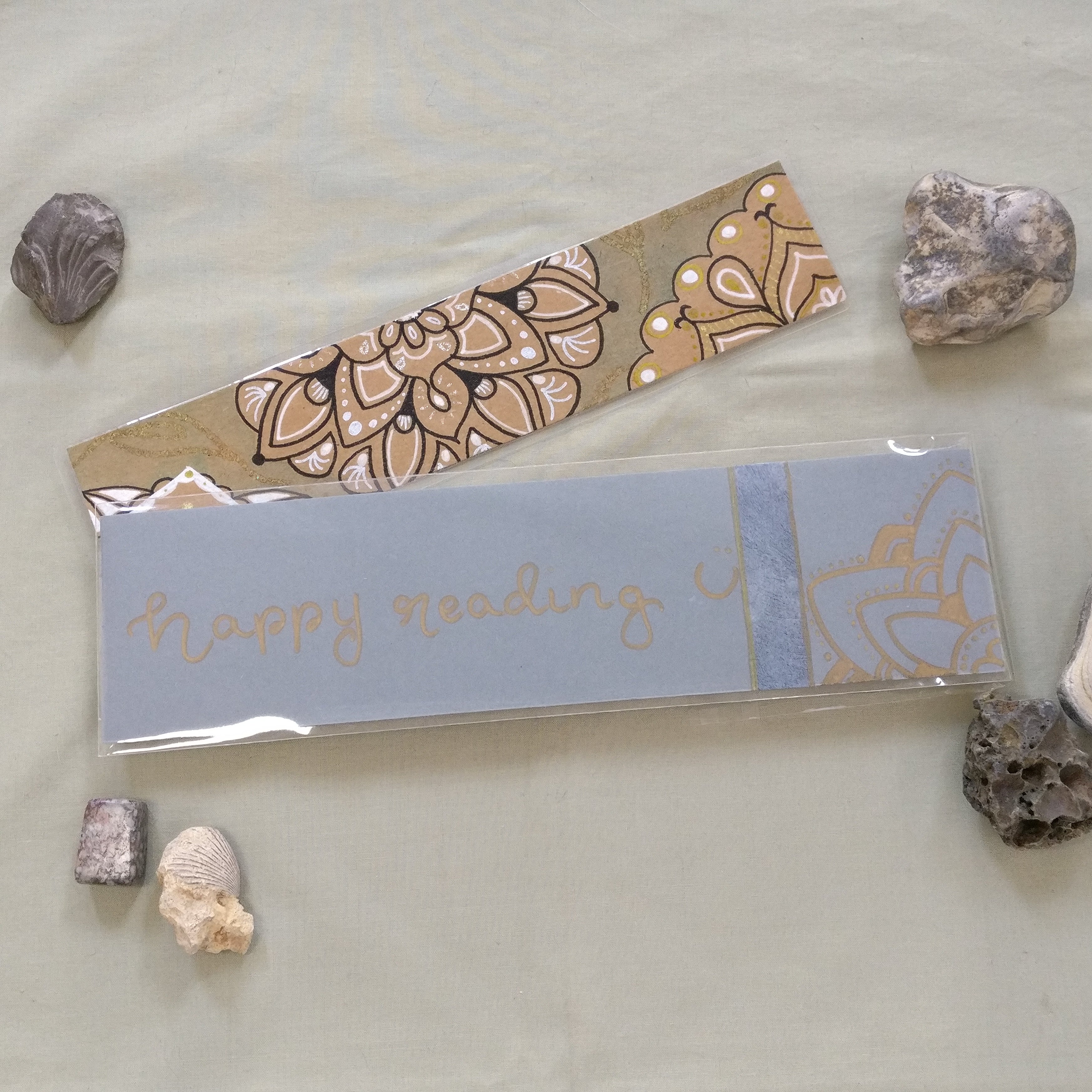 An aqua-coloured Mandala Bookmark with one of the handmade package backings with a hand-written "happy reading" and a mandala corner in gold.