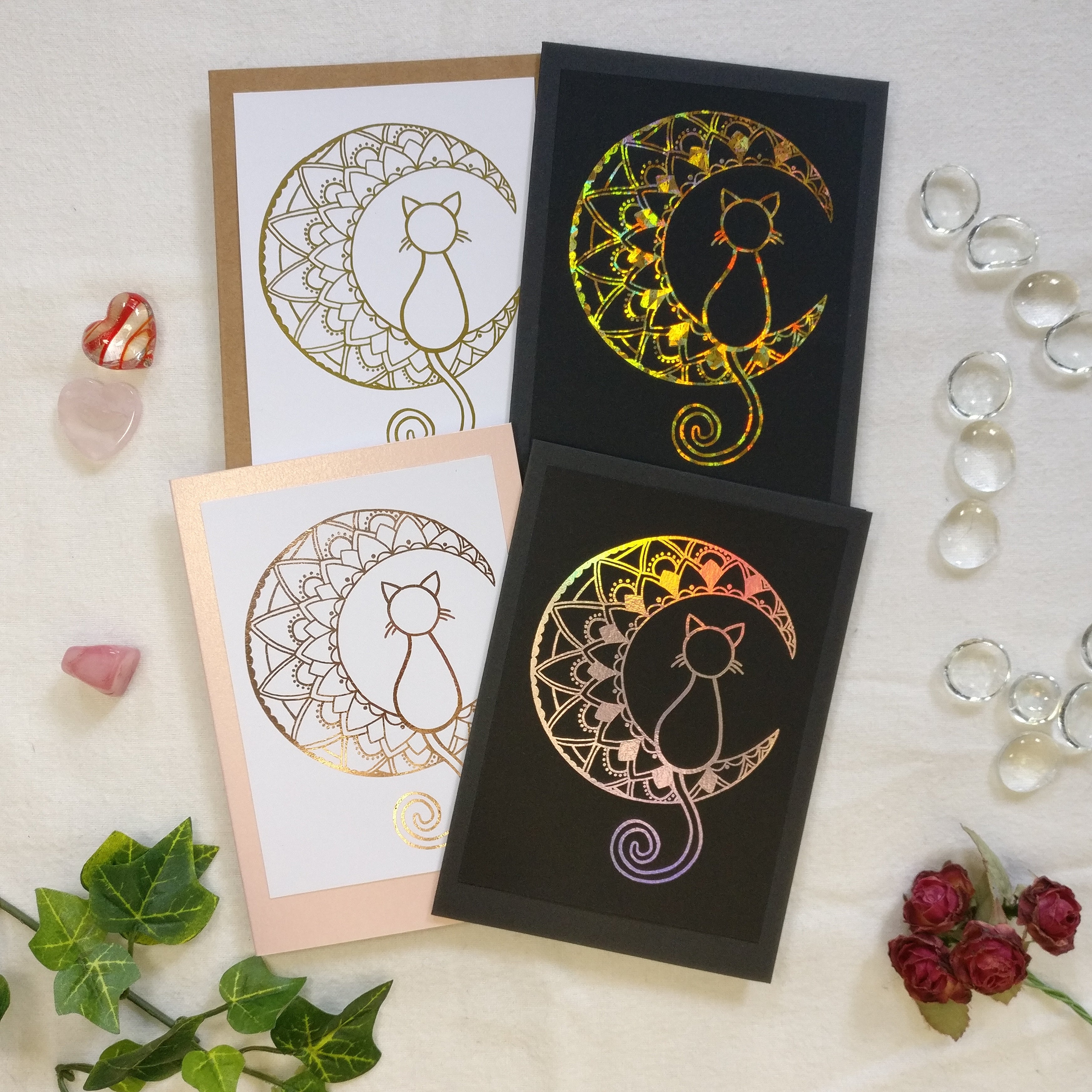 Handmade cards with a cat sitting in a crescent mandala moon. Four cards showing cardstock colours and foild options. 
Top row: Gold foil on white and kraftpaper card and Holographic crystal gold foil on black card. 
Bottom row: Rose-gold foil on white and pearly pink and holographic silver foil on black card.
