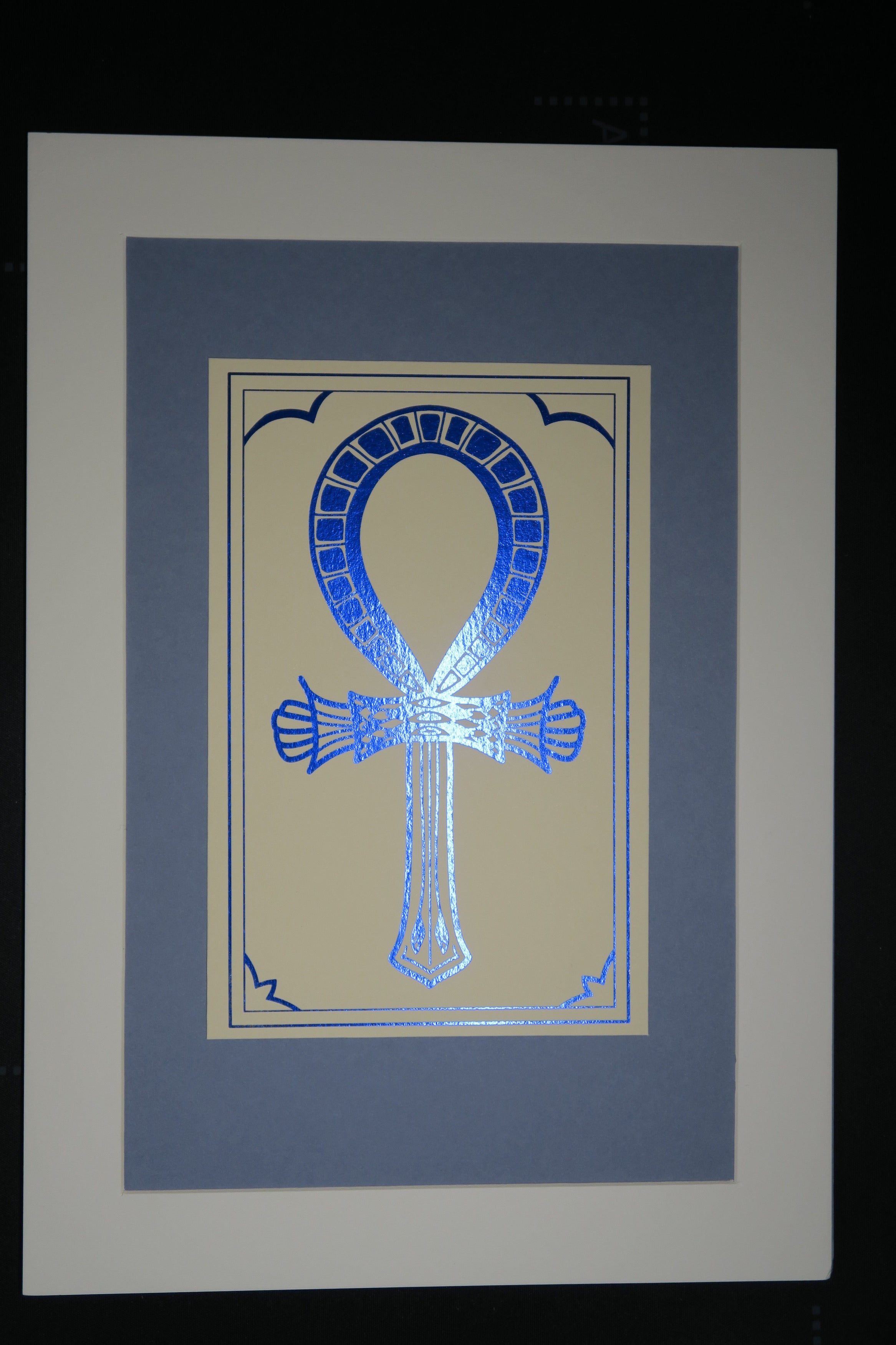 The blue foiled Ankh symbol mounted on medium grey backing.