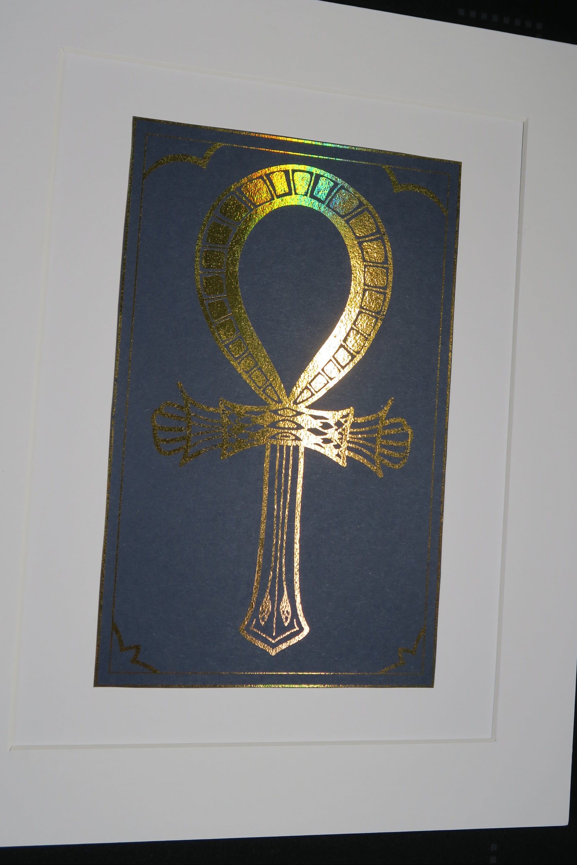 Holographic Gold foiled Ankh symbol on a navy blue card mounted on white backing.