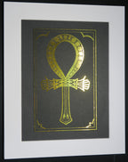 Gold foiled ankh symbol on black card mounted on white backing.
