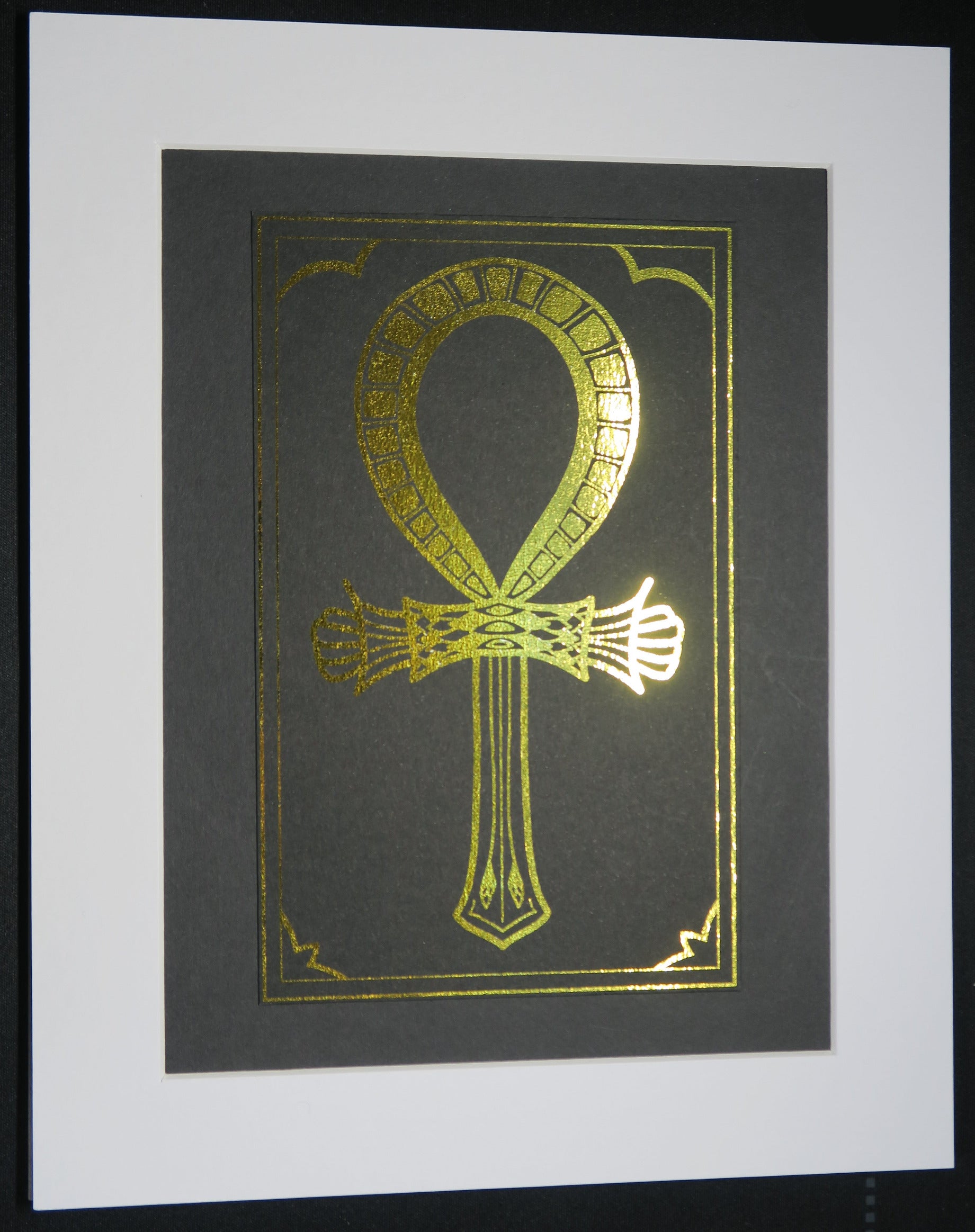 Gold foiled ankh symbol on black card mounted on white backing.