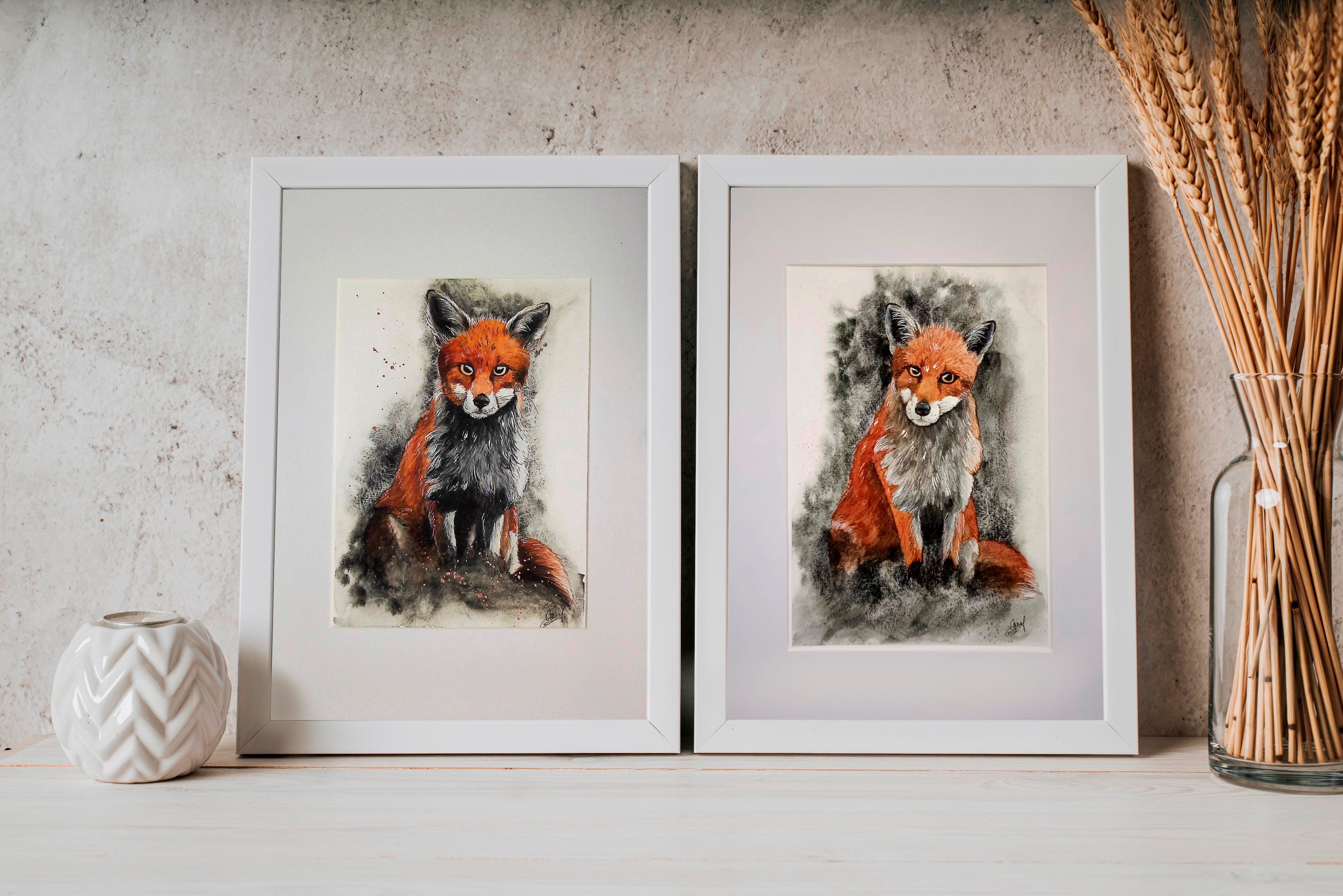 Two framed artworks of foxes on a neutral background with decorative elements. The two fox versions available. On the left is Fox Version 1, on the right is Fox Version 2 (which has slightly lighter-coloured chest fur.
