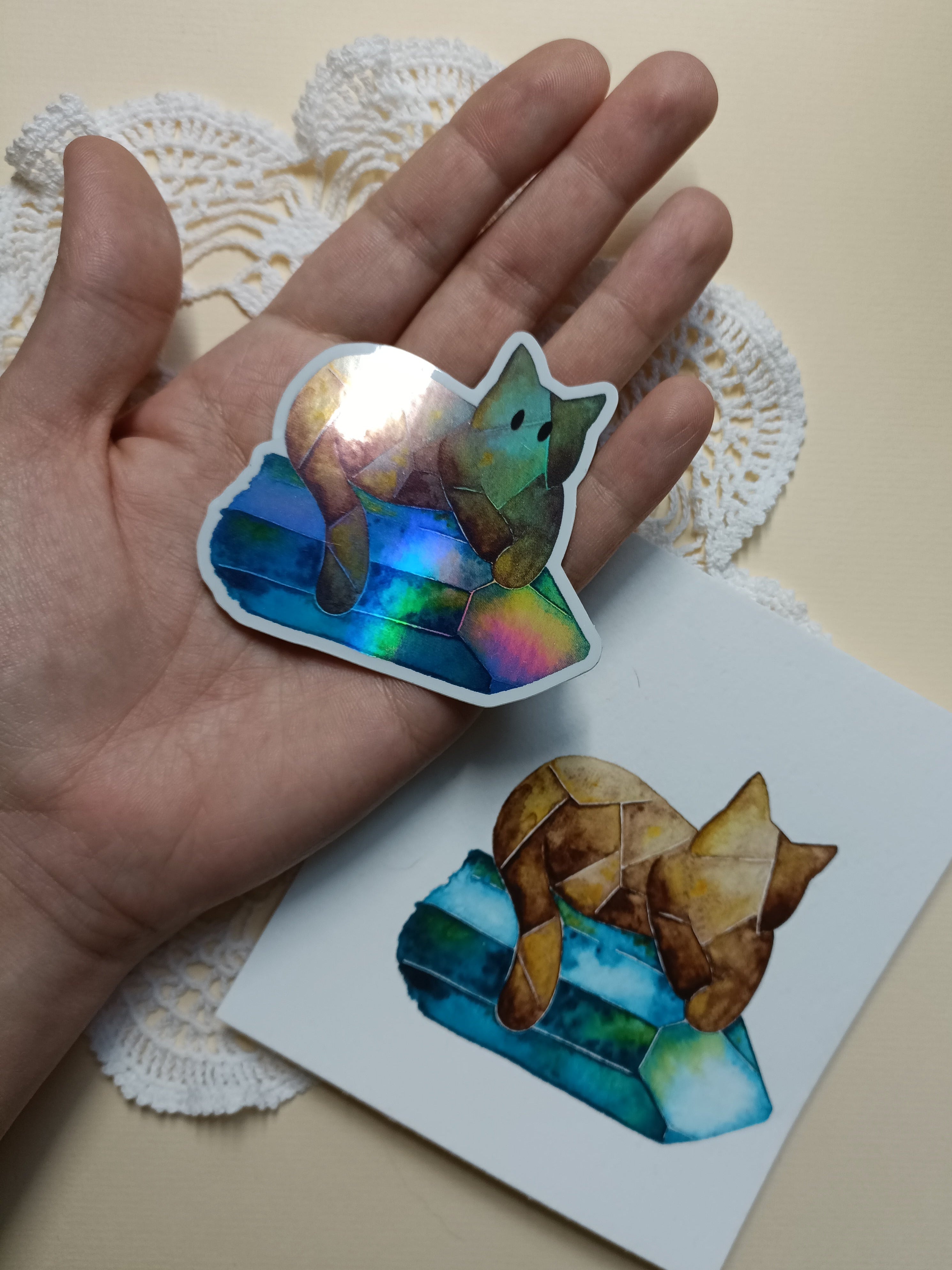 Hand holding a holographic cat crystal sticker for size reference, next to the original watercolor illustration.