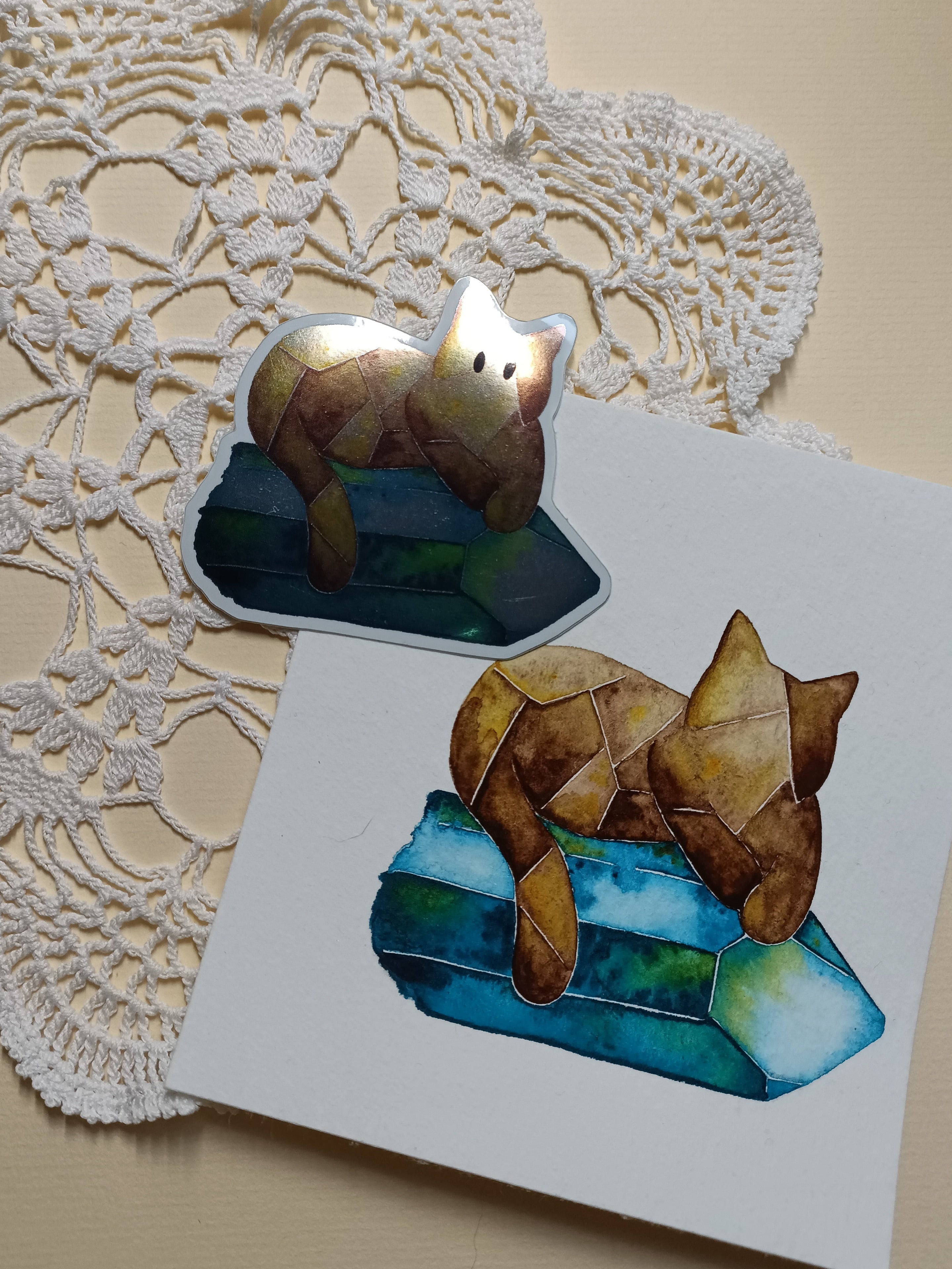 Mirror crystal cat sticker next to original watercolour painting, layed out on a light-coloured background.